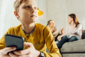 A child uses their smartphone while their parents talk on the sofa in the background.